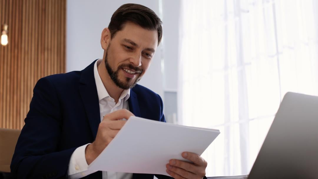 Man in business attire looking at a paper at his desk in front of his laptop