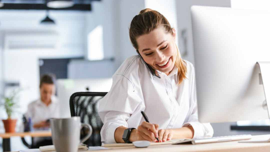 Businesswoman talking on the phone at her desk in front of a laptop taking notes