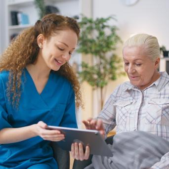 woman in a blue scrub top sitting and talking with an elderly woman pointing to a tablet